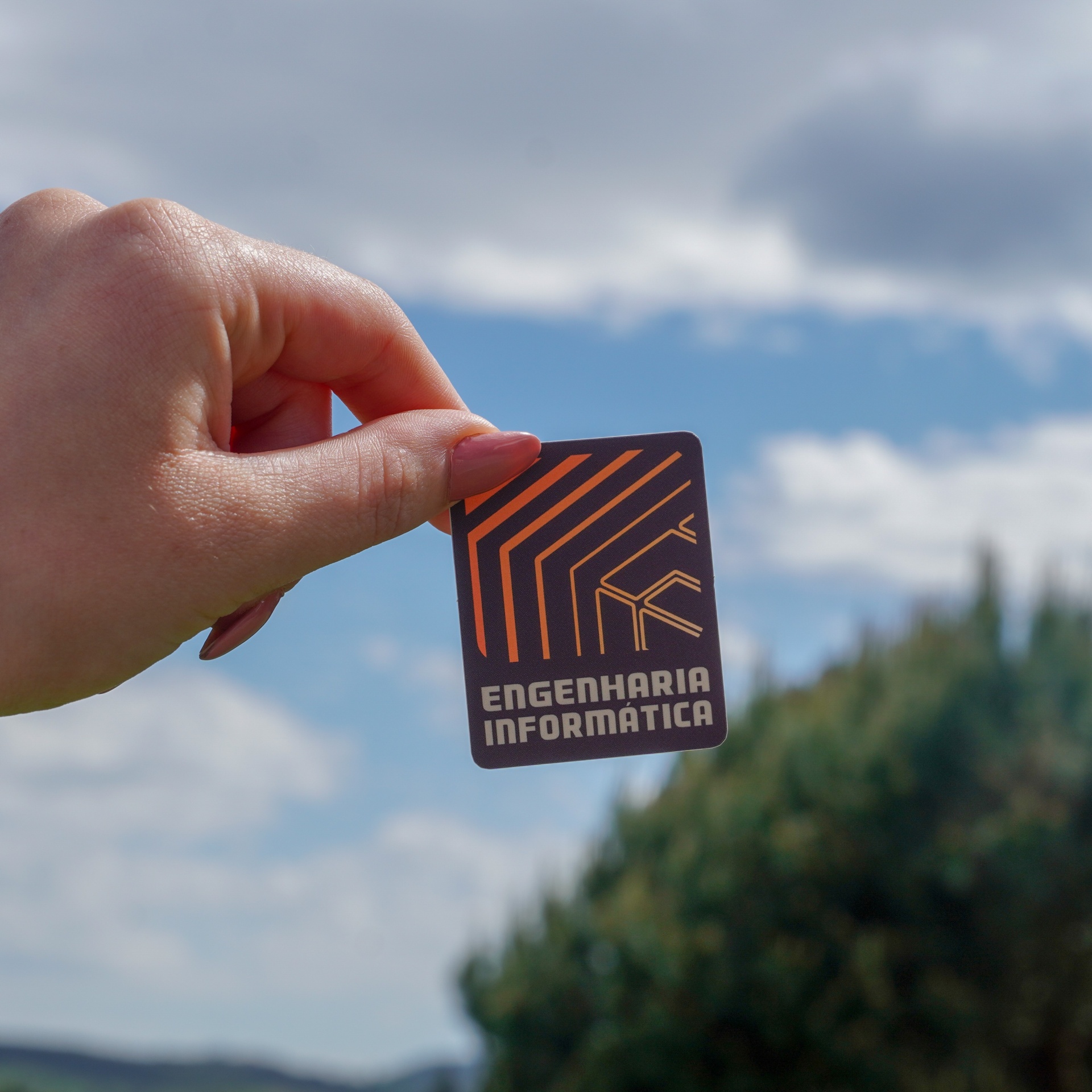 Cartão preto com linhas laranja e texto ENGENHARIA INFORMÁTICA segurado por mão em fundo de céu e árvores