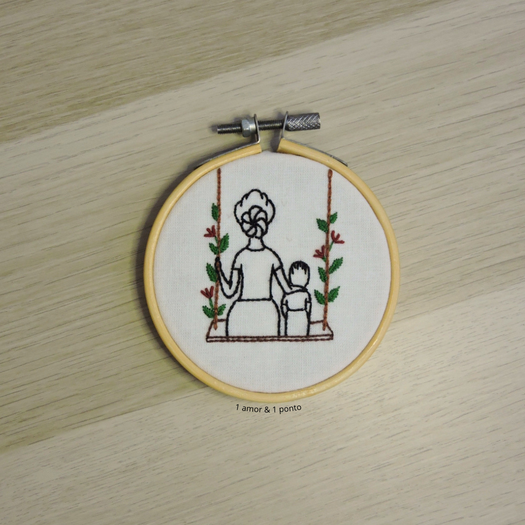 Aro de bordado com tecido branco e bordado de mulher e criança num baloiço