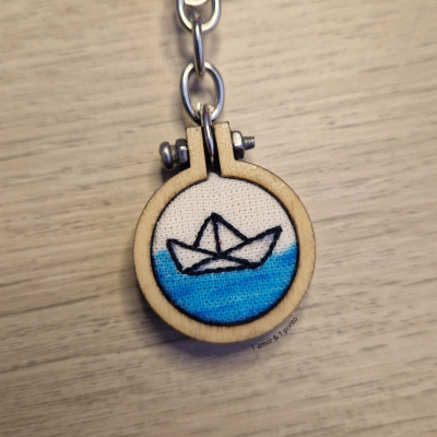 Chaveiro com bordado de barco de papel em fundo azul em aro de madeira sobre superfície clara