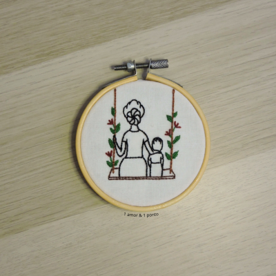 Aro de bordado com tecido branco e bordado de mulher e criança num baloiço