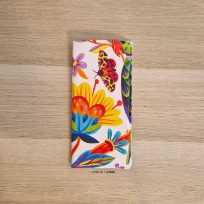 Carteira larga e alta em tecido colorido com padrão de flores e borboleta sobre mesa de madeira.