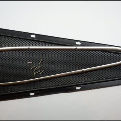 Grelha preta trapezoidal com textura perfurada, marco metálico prateado e parafusos.