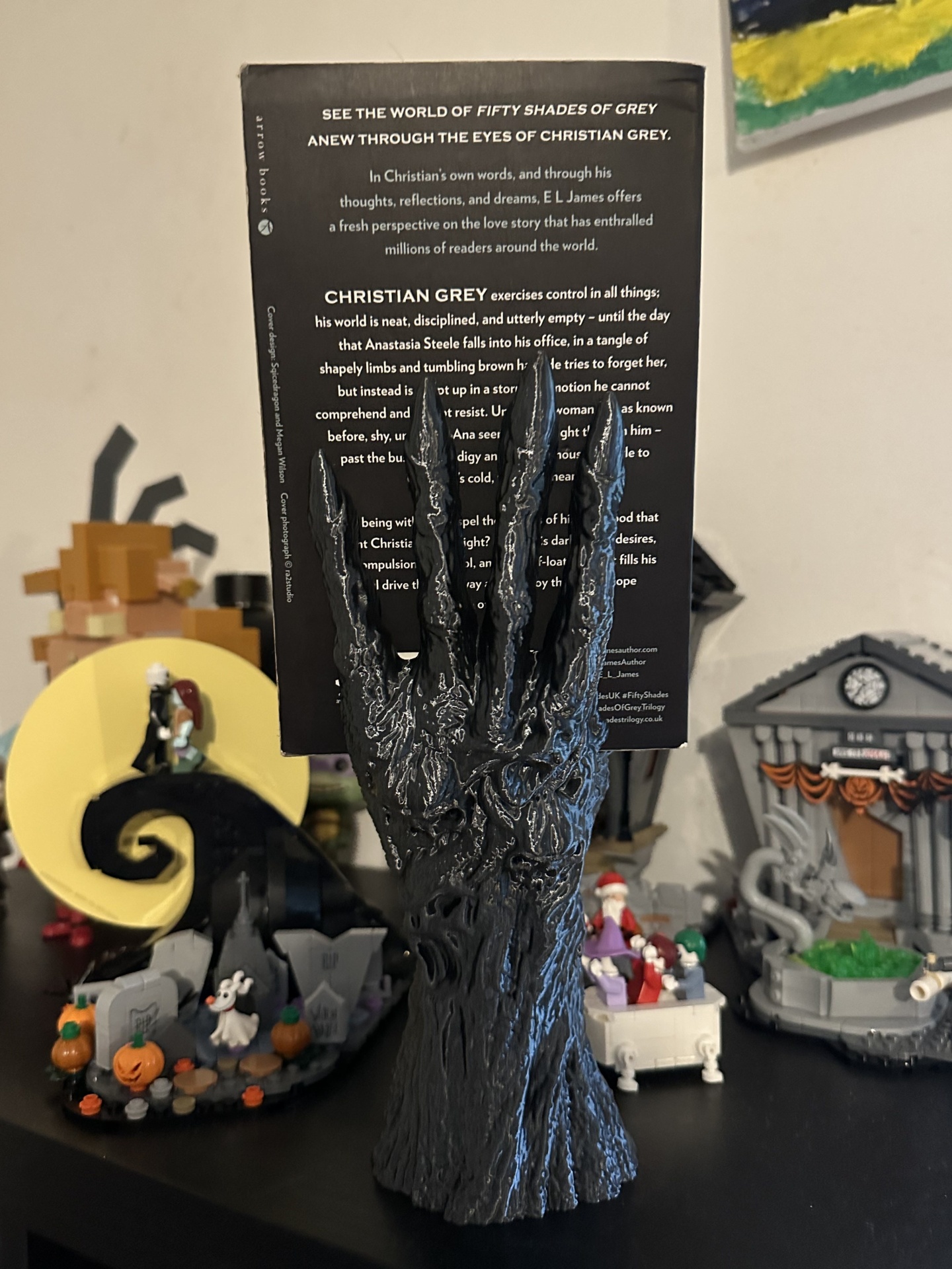 Mão escultórica preta segurando livro Fifty Shades of Grey com decoração de Halloween ao fundo