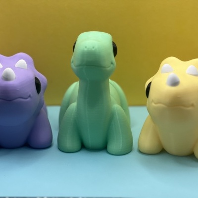 Três brinquedos de dinossauros coloridos em plástico ou borracha sobre superfície azul com fundo amarelo