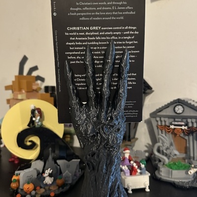 Mão escultórica preta segurando livro Fifty Shades of Grey com decoração de Halloween ao fundo