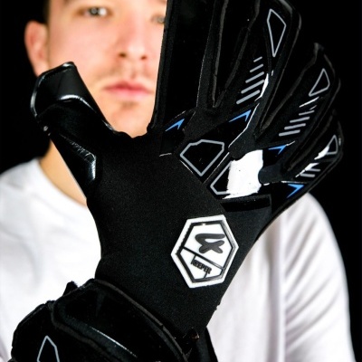 Luvas de guarda-redes pretas com detalhes brancos e azuis em fundo preto e pessoa ao fundo