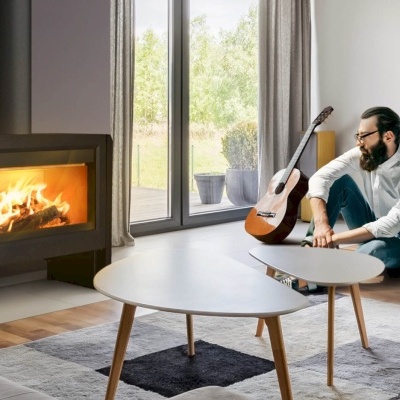 Lareira moderna preta acesa numa sala com mesa branca, tapete cinzento e homem sentado com guitarra