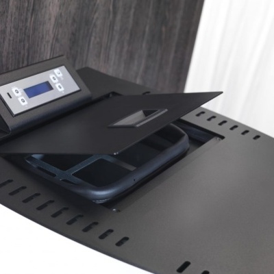 Mesa preta com painel digital e ventilação numa sala com paredes cinzentas e cortina branca