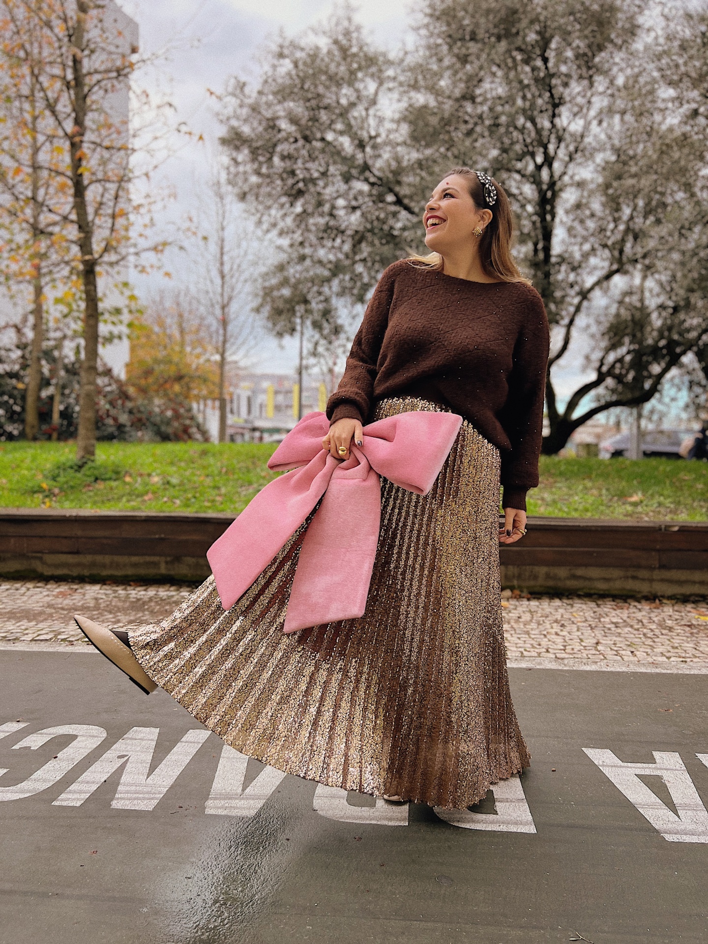 SAIA NAYARA Mulher com camisola castanha, saia dourada plissada e clutch rosa com laço grande no parque.