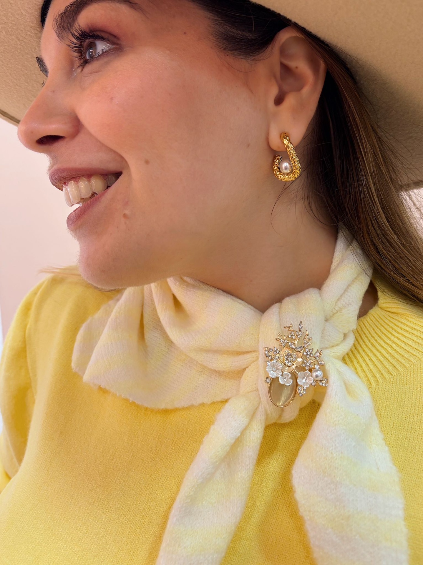 PREGADEIRA PATROCÍNIA Mulher usando brincos dourados com pérola e cachecol amarelo claro com broche dourado com pérolas em fundo claro