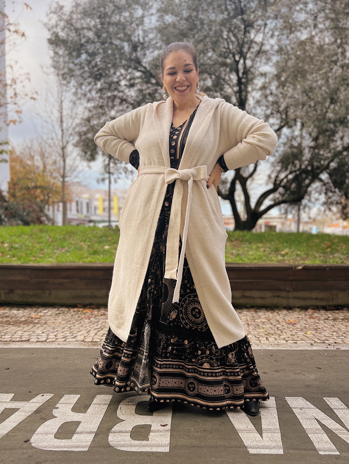 CASACO NATALI Mulher sorridente com casaco creme e vestido preto com padrão celestial, ao ar livre