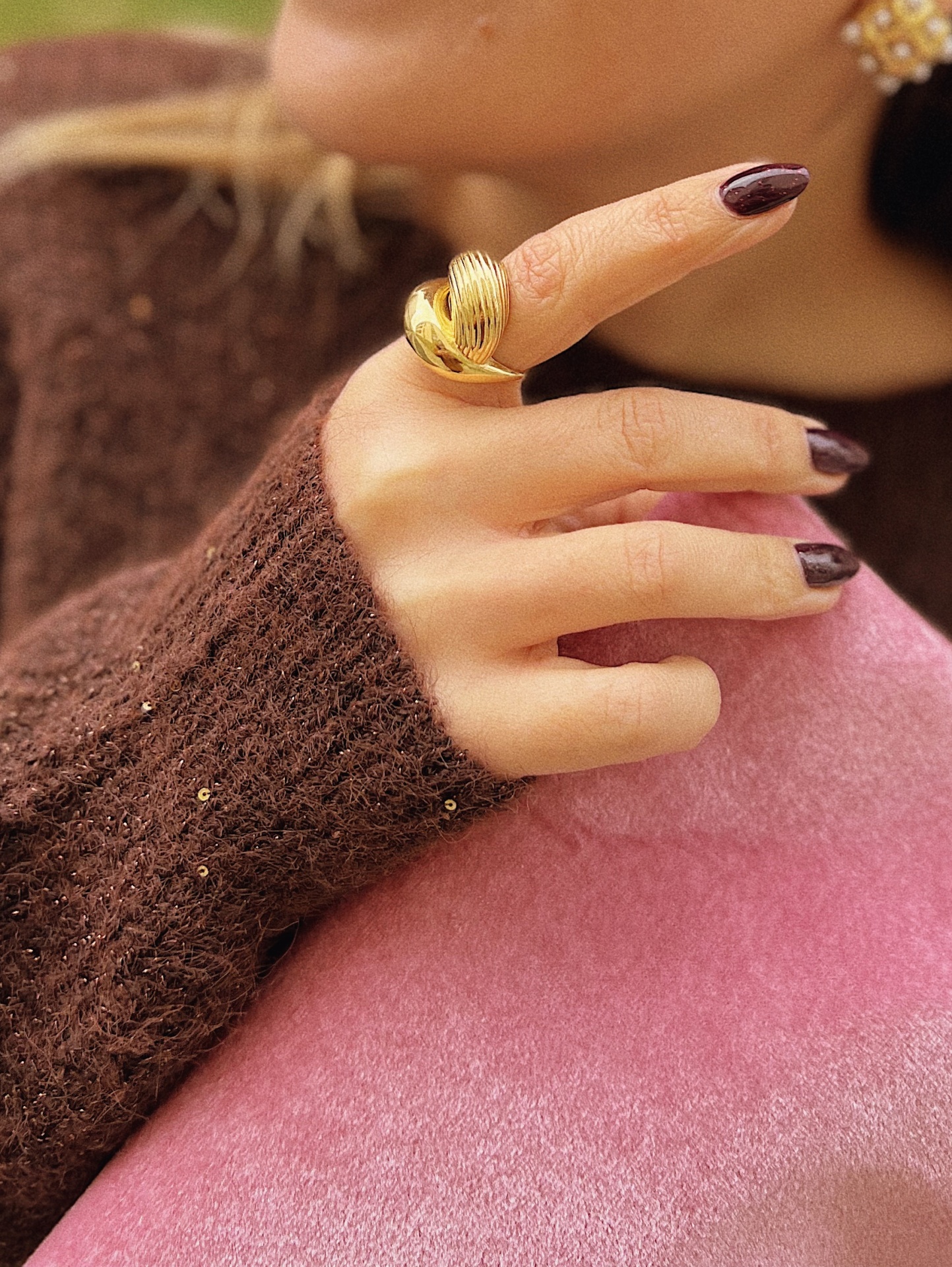 ANEL NÉRCIA Mão com anel dourado e camisola castanha com lantejoulas sobre tecido rosa.