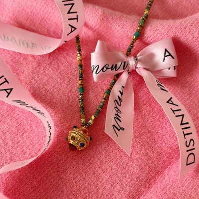 Colar com contas verdes e dourado, pendente dourado com pedras coloridas, fita rosa com texto em preto sobre tecido rosa.