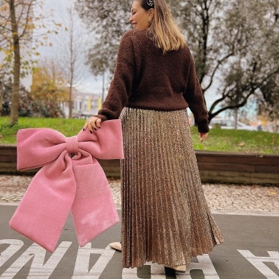 Mulher com camisola castanha, saia dourada plissada e grande laço rosa segurado ao lado, em rua com árvores ao fundo.
