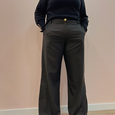 Conjunto feminino com camisa preta de botões dourados e calças cinzentas com riscas finas, sapatos vermelhos, parede rosa e chão de madeira.