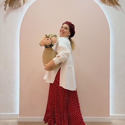 Mulher com camisa branca, saia vermelha de bolinhas, boina vinho, segurando vaso com flores cor de rosa