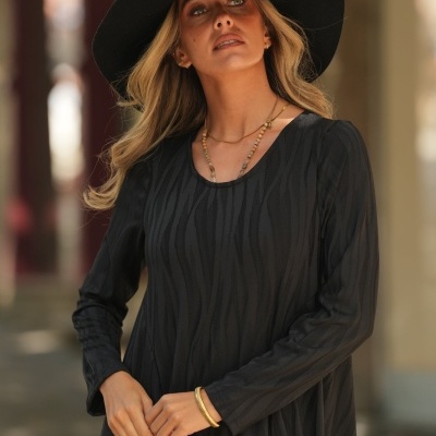 Mulher de vestido preto comprido com padrão, chapéu preto de aba larga e acessórios dourados.