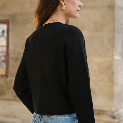 Mulher de perfil e costas com camisola preta de manga comprida e jeans azuis, usando brincos dourados, junto a parede de pedra clara