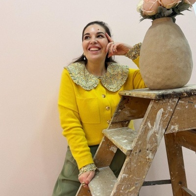 Mulher com blusa amarela e gola floral, calças verdes, ao lado de escada de madeira com vaso atopado de flores cor-de-rosa.