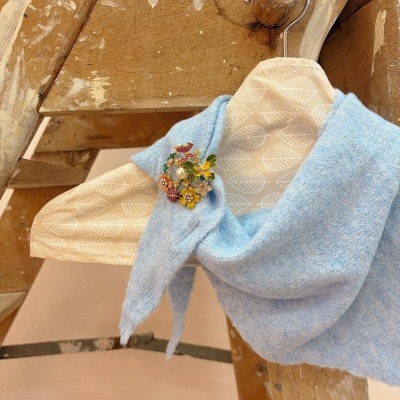 Cachecol azul claro com broche floral preso em cabide de madeira com padrão de folhas, pendurado em escada de madeira.