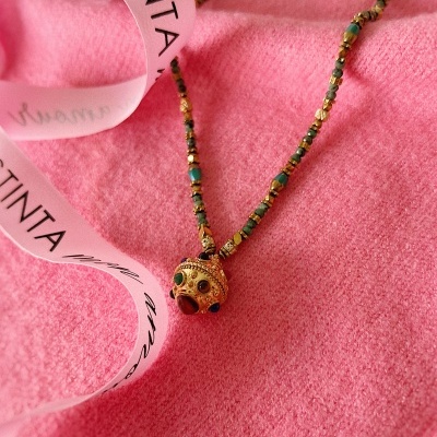 Colar dourado com contas verdes e pendente sobre tecido rosa e fita branca com texto