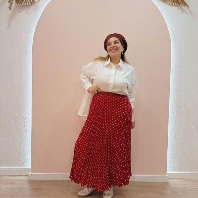 Mulher com camisa branca, saia vermelha com bolinhas brancas, boina vermelha e sapatilhas claras em parede rosa com arcada