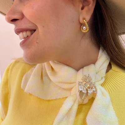Mulher usando brincos dourados com pérola e cachecol amarelo claro com broche dourado com pérolas em fundo claro