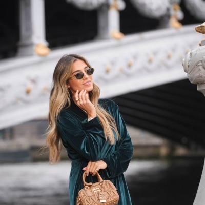 Mulher com vestido verde de veludo e mala bege MIU MIU perto de ponte decorada