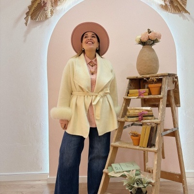 Mulher com casaco creme e chapéu rosa junto a escada de madeira com livros e vasos