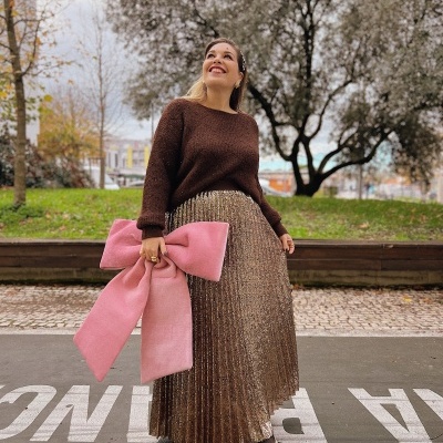 Mulher com camisola castanha, saia dourada plissada e bolsa rosa em laço num parque.