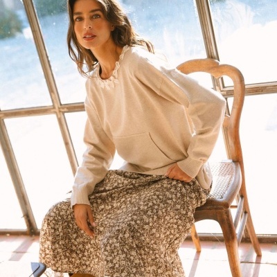 Mulher sentada com camisola bege e saia floral sentada junto a janela