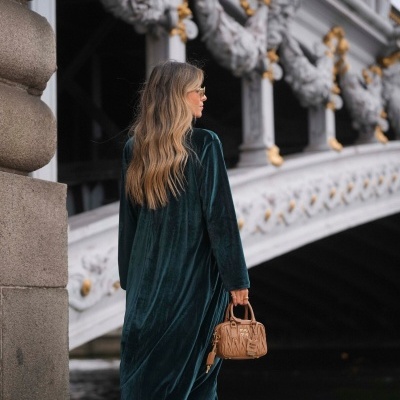 Mulher com vestido verde veludo, botas castanhas e mala MIU MIU castanha junto a ponte ornamentada