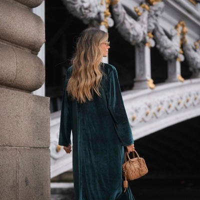 Mulher com vestido verde de veludo e mala bege junto a estrutura branca decorada.