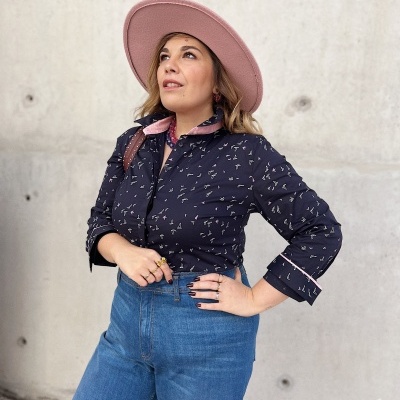 Mulher com camisa preta com padrão e chapéu rosa junto a parede cinza