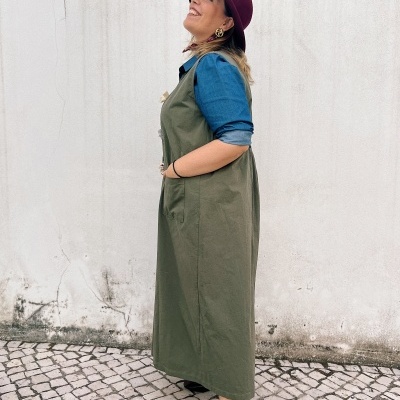 Mulher de perfil com vestido verde, camisa azul, chapéu vinho e sapatos vinho, em calçada portuguesa e parede branca