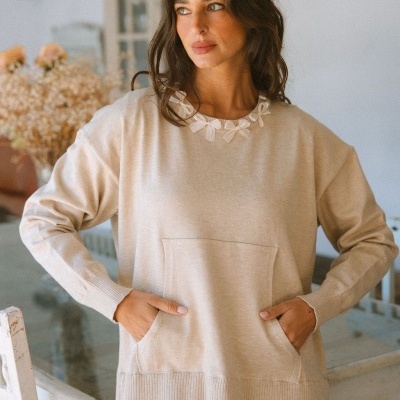 Casaco bege com bolso frontal e gola decorada, usado por mulher num ambiente interior.