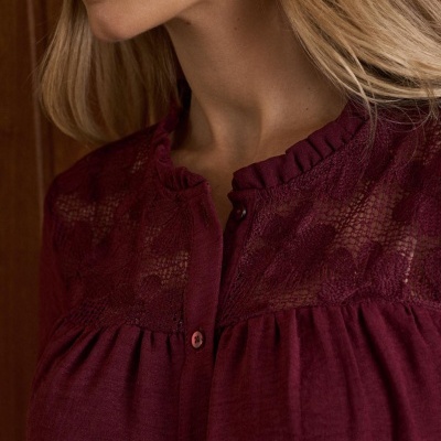 Blusa vinho com detalhes rendados e botões, vista parcial de mulher com cabelo loiro e fundo de madeira