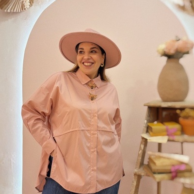 Mulher com chapéu e camisa rosa claro e calças de ganga azuis, em ambiente com arco e prateleira com livros e vasos.
