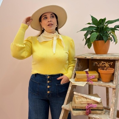 Mulher com chapéu bege, camisola amarela e jeans escuros junto a escada com livros e vasos