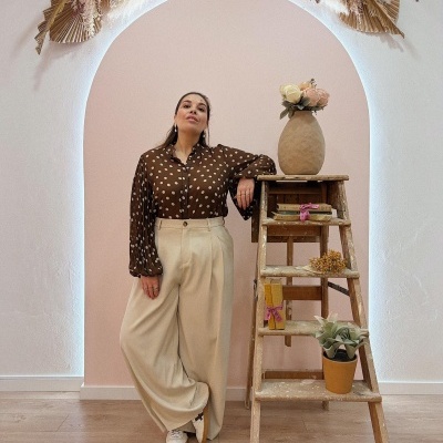 Mulher com blusa castanha às flores e calças creme, escada de madeira com livros e plantas, fundo rosa com arco decorado