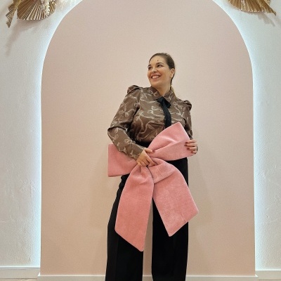 Mulher com blusa castanha e calças pretas segurando bolsa rosa grande laço junto a arco decorativo.