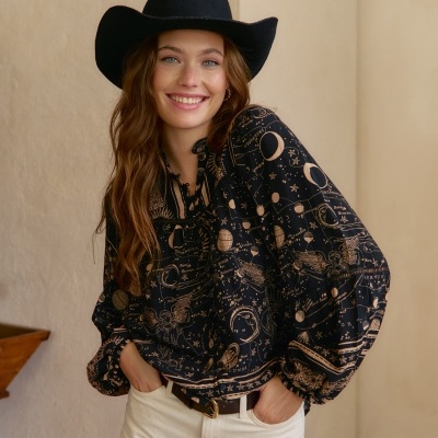 Mulher com blusa preta de mapas astrológicos bege, calças brancas e chapéu preto