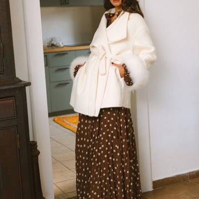 Mulher com casaco branco felpudo com cinto e punhos de pelo sobre vestido castanho de bolinhas brancas em interior.