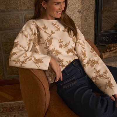 Mulher com camisola creme de flores castanhas e calças de ganga azul sentada numa cadeira castanha junto a uma lareira.