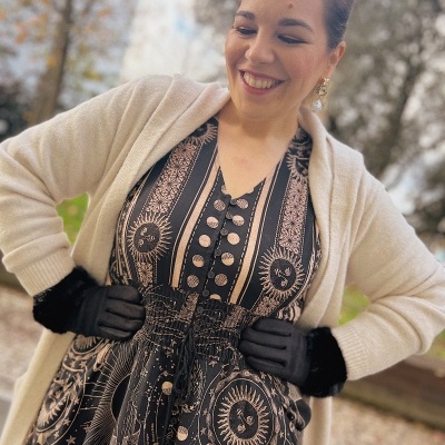 Mulher de sorriso vestindo vestido com padrão celeste, cardigan bege e luvas pretas.