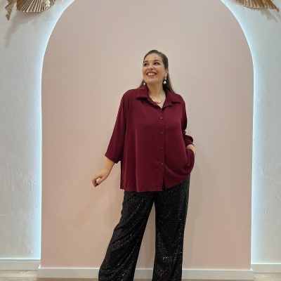 Mulher vestindo camisa borgonha e calças pretas de lantejoulas em fundo rosa com arco decorado