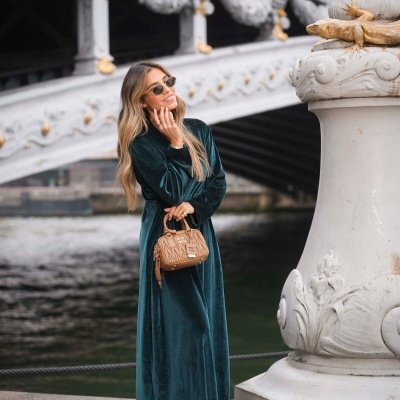 Mulher com vestido verde escuro de veludo, óculos de sol e mala castanha em ambiente urbano junto a ponte e pilar decorativo