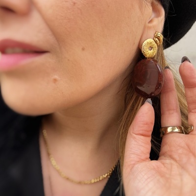 Brincos dourados com pedra castanha em orelha de mulher com chapéu preto.