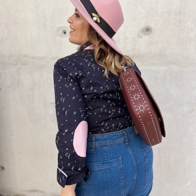 Mulher de lado com chapéu rosa, camisa azul com cotoveleiras rosas, calças de ganga e mochila castanha perfurada