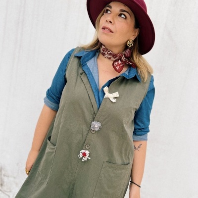 Mulher com chapéu bordô, camisa azul, macacão verde com broches e lenço vermelho e branco no pescoço.